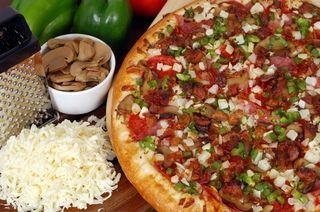 Pizza with toppings, ingredients on the side: mushrooms, cheese, green peppers, on wooden surface.