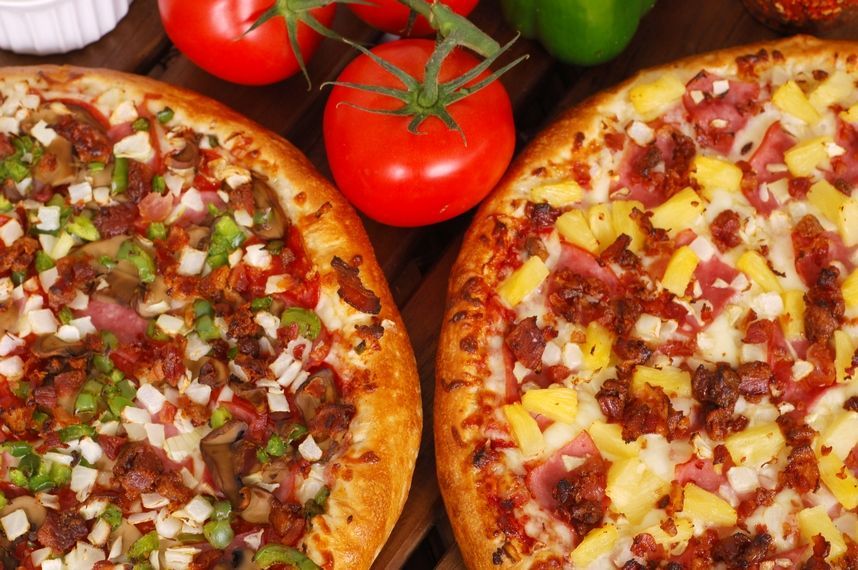 Two pizzas are sitting on a wooden table next to tomatoes and peppers.