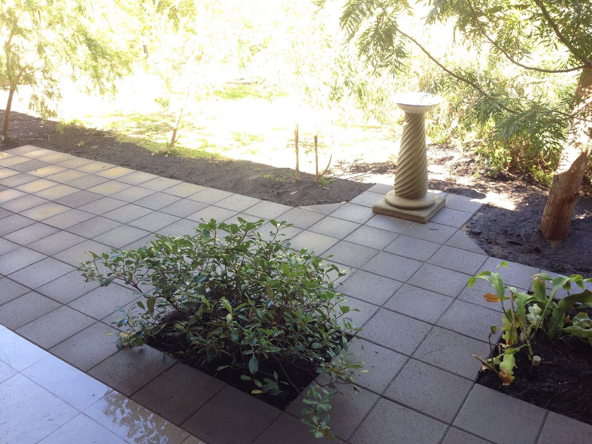Patio with square gray tiles, small bush, decorative column, and sunny garden background.