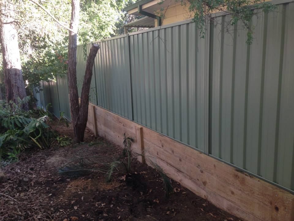 Green metal fence with wooden base, along a yard with trees and dirt.