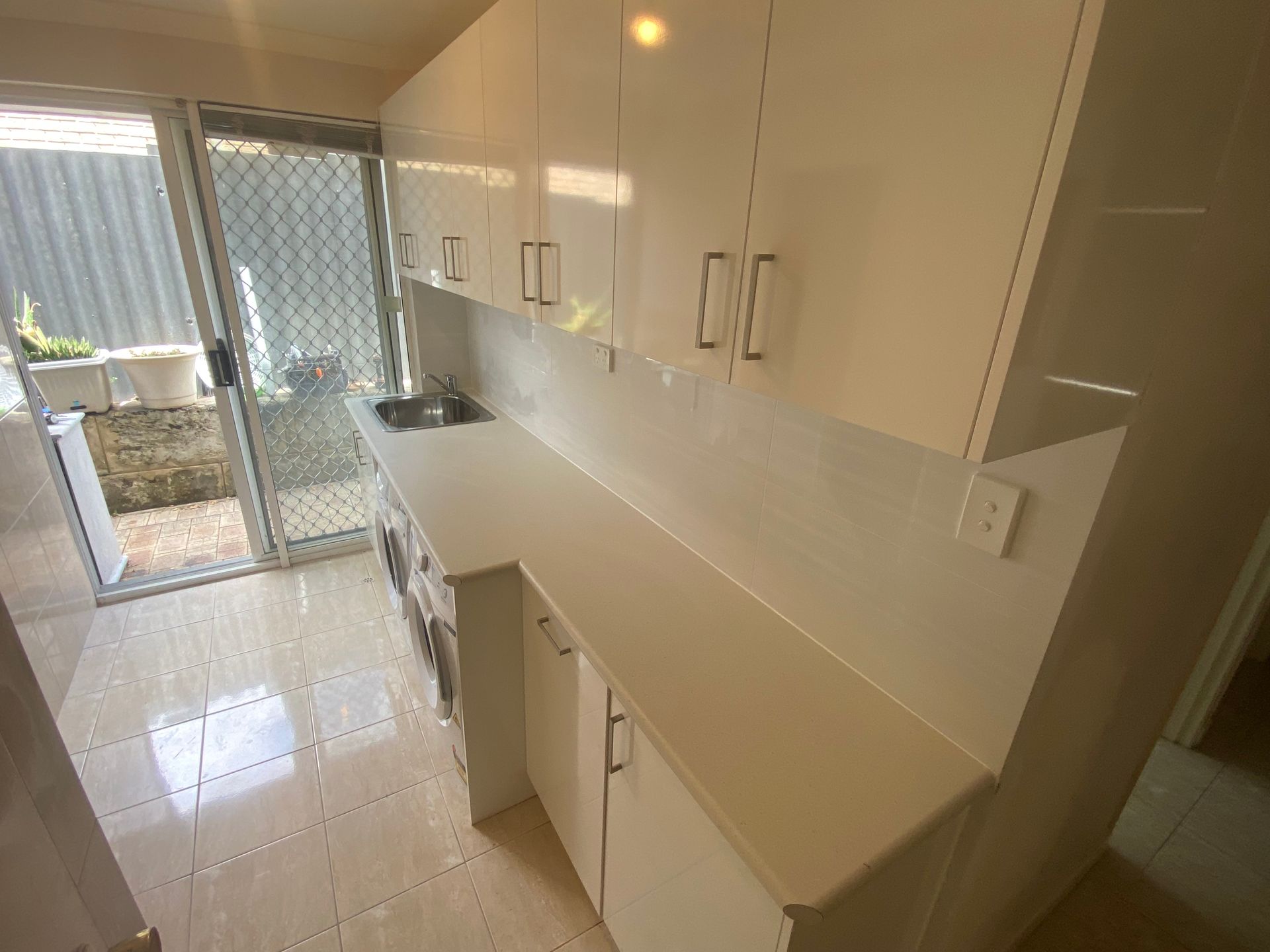 White cabinets and countertops in a small laundry room with a sink, and a sliding glass door.