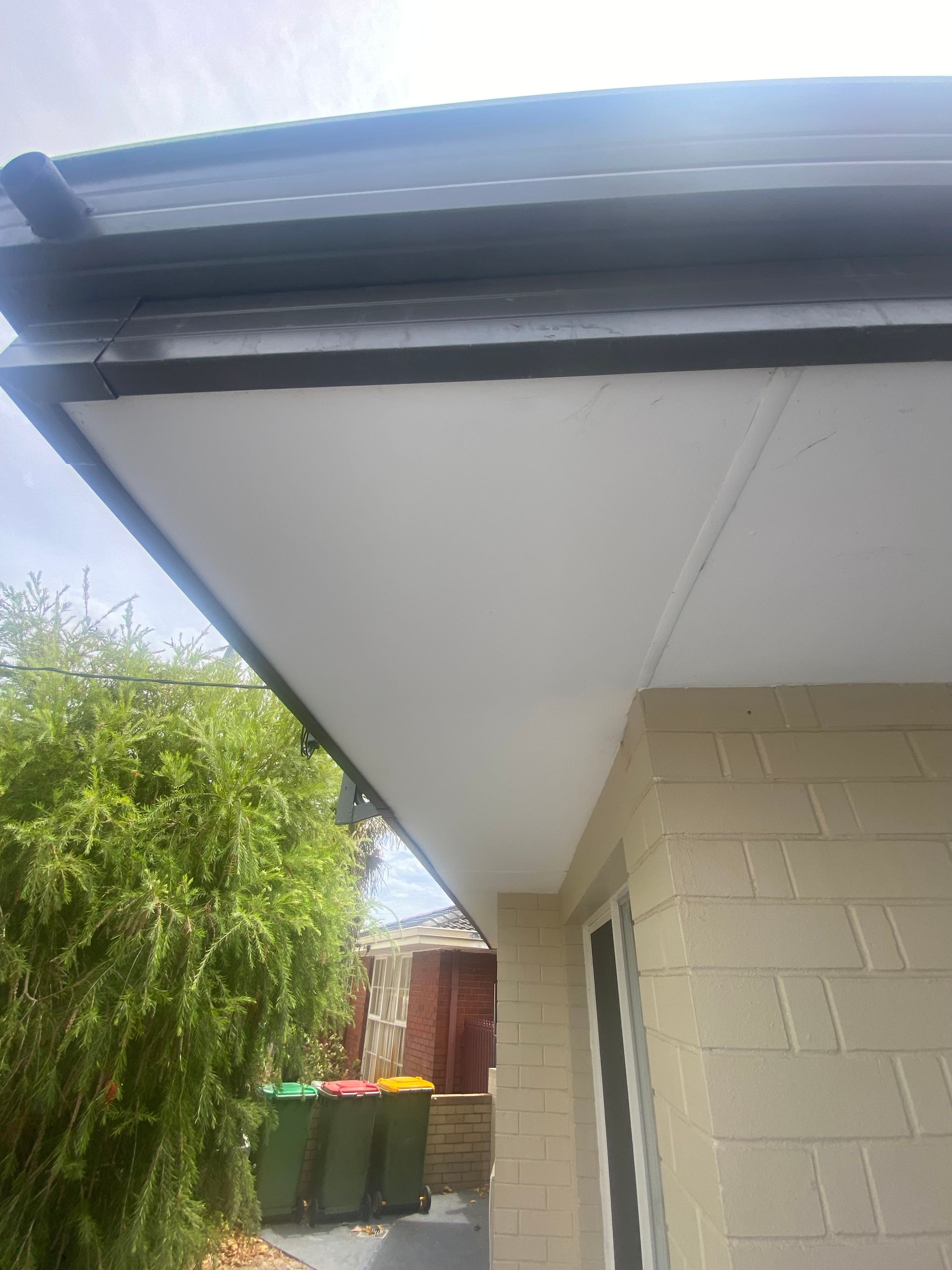 A white soffit and black gutter of a house, with a brick wall and green bins visible.