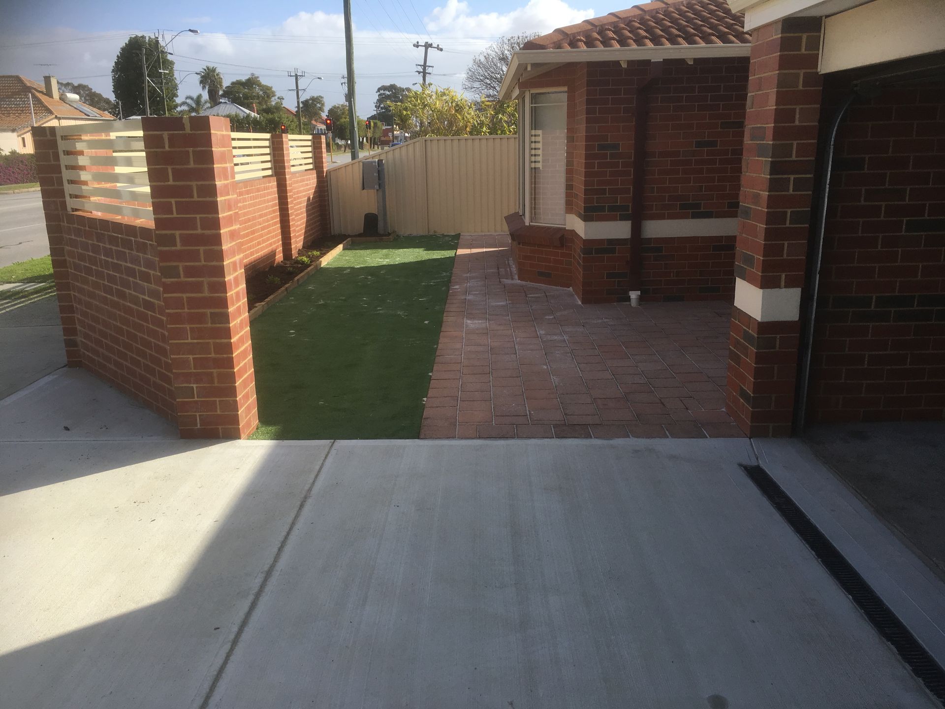 Brickwork entrance with a grass patch and brick path, bordered by a concrete driveway and fence.