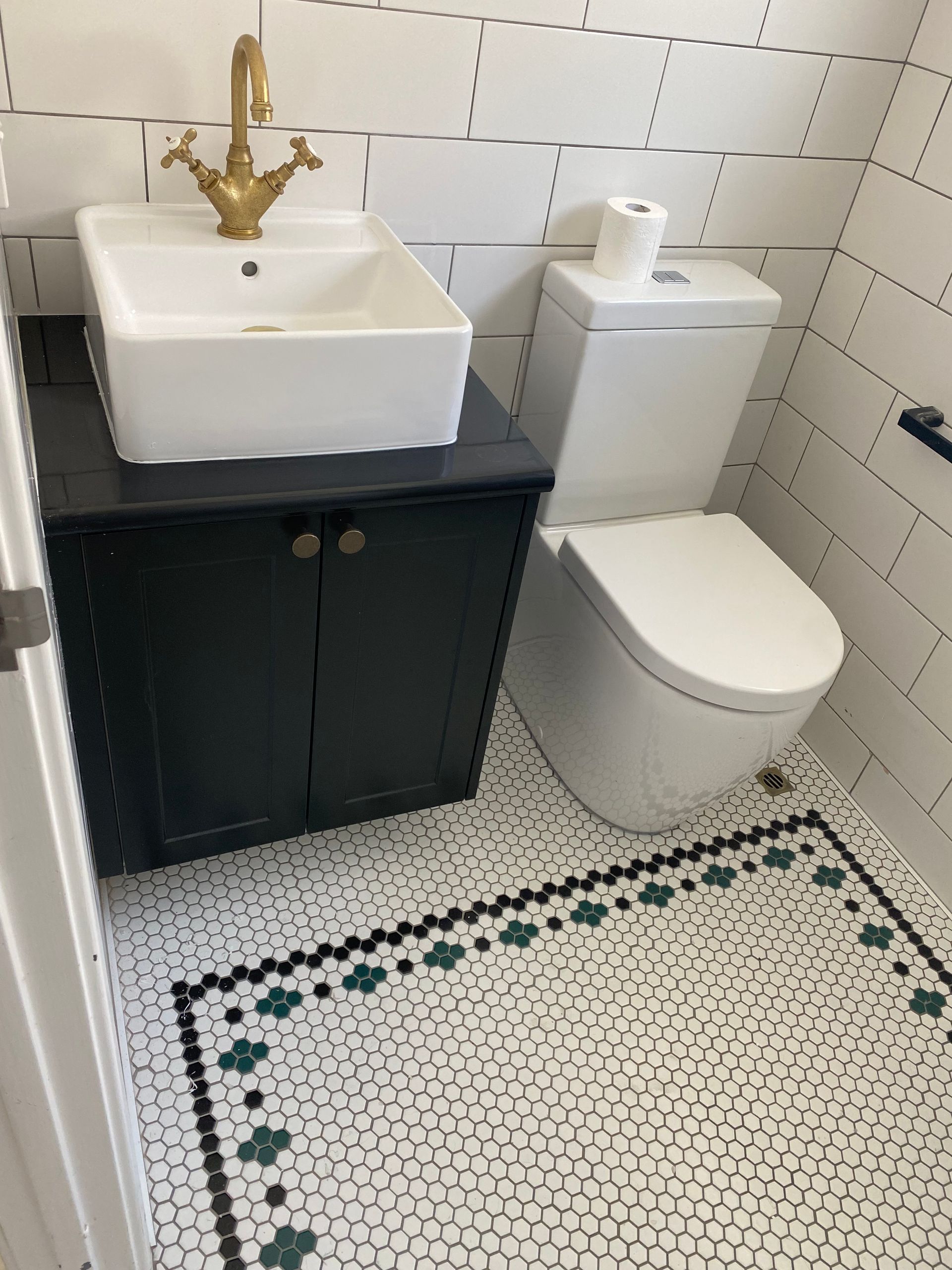 Bathroom with dark blue vanity, white sink, gold faucet, white toilet, and patterned floor. White subway tile walls.