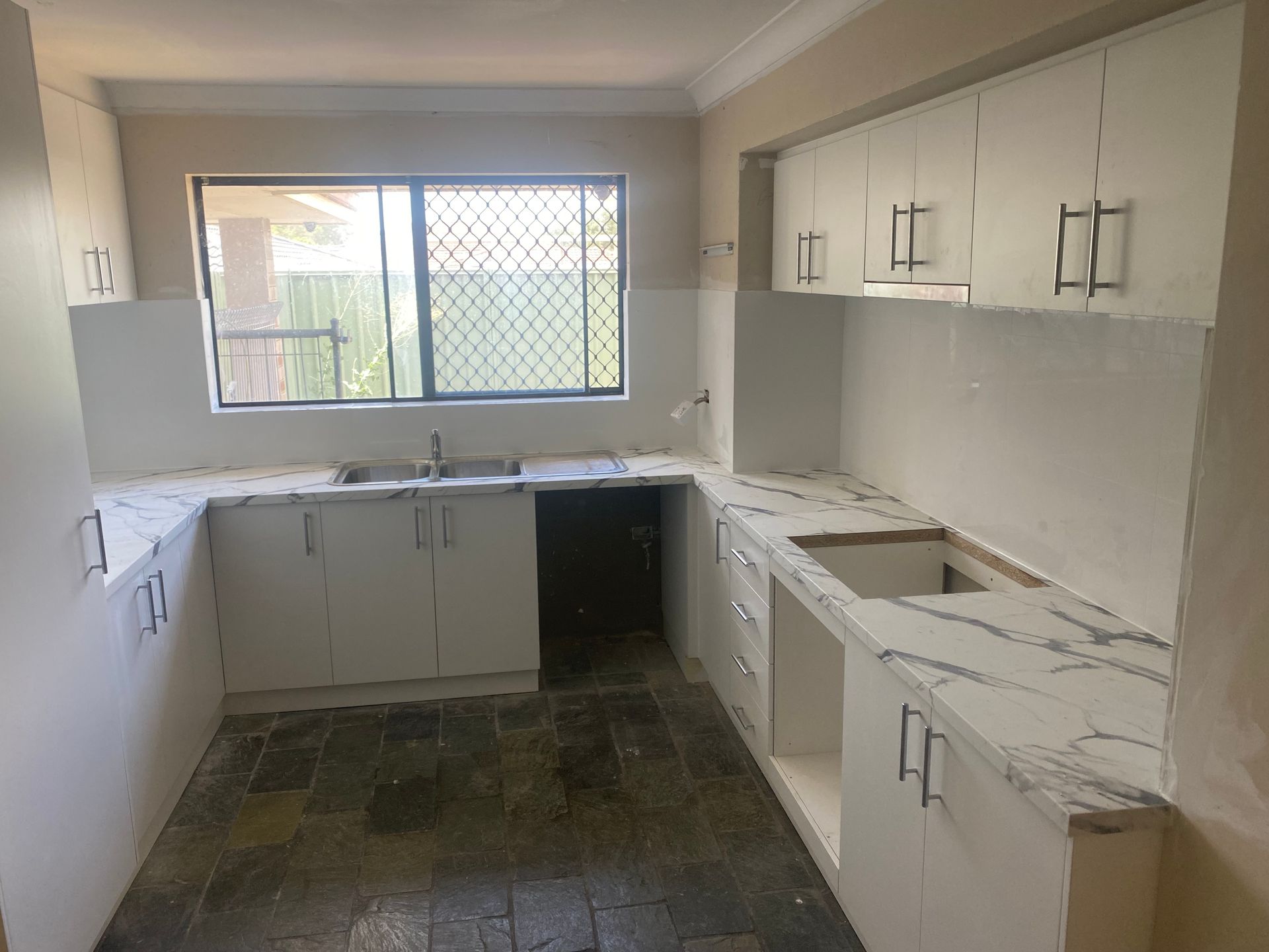 Empty kitchen with white cabinets, marble-look countertops, and a window.