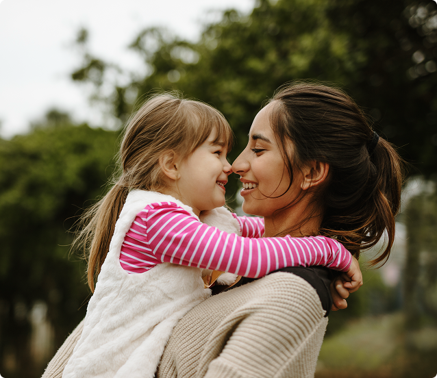 Woman holding and smiling at a child, touching noses. Outdoors with trees in the background.