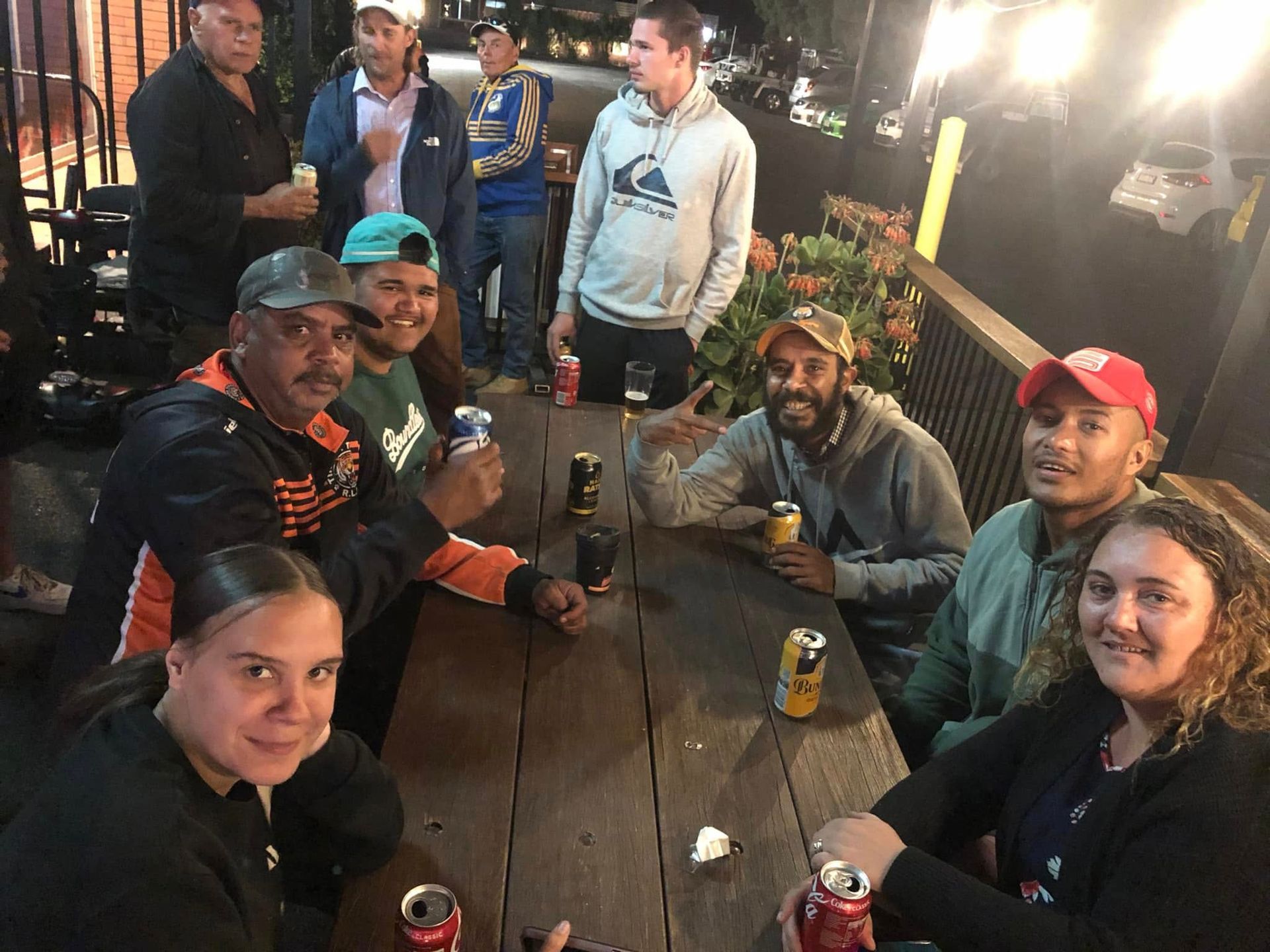 A group of people are sitting around a wooden table holding cans of soda.