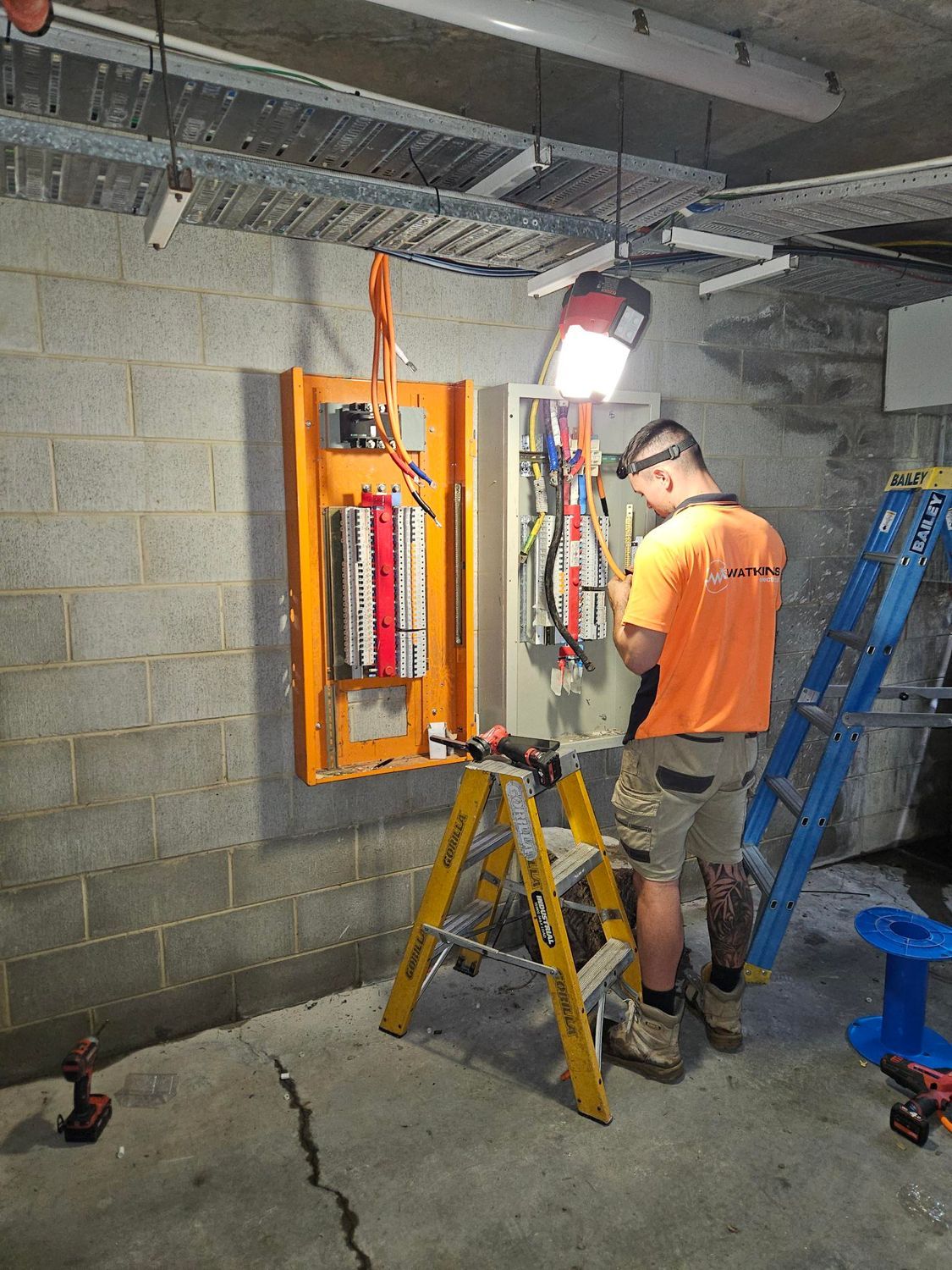Electrician in orange shirt works on electrical panel.  Tools on a ladder, in a concrete room — Watkins Electrical In Pialba, QLD