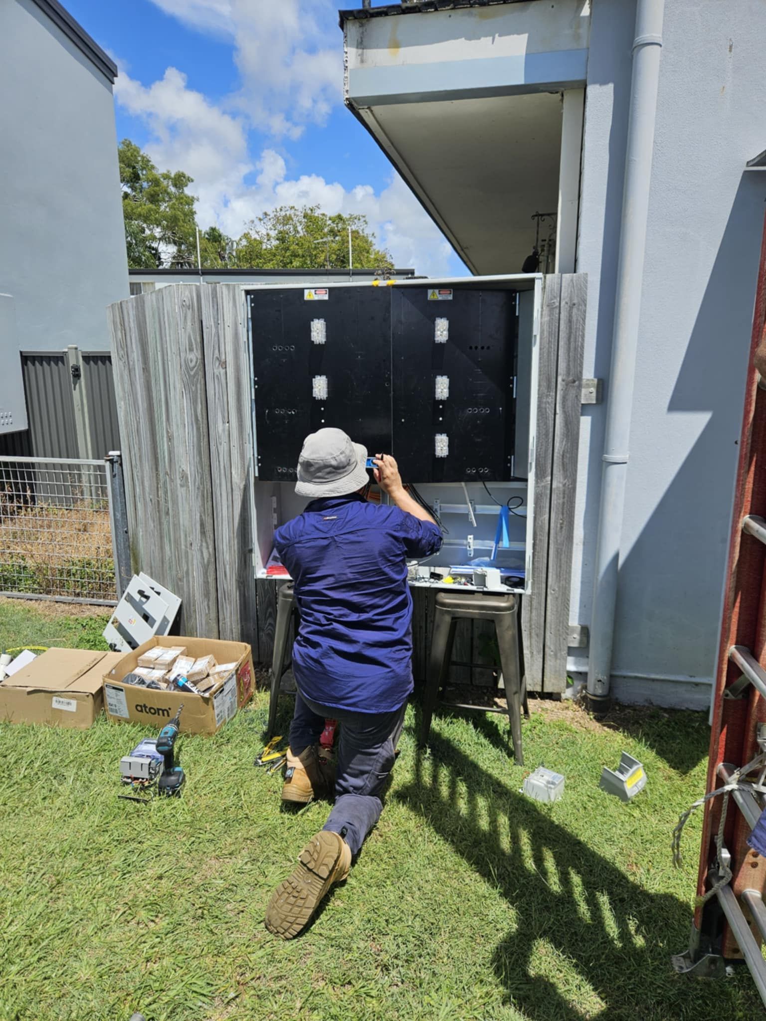 Person working on an electrical panel outdoors. Sunny day, green grass, tools on the ground.