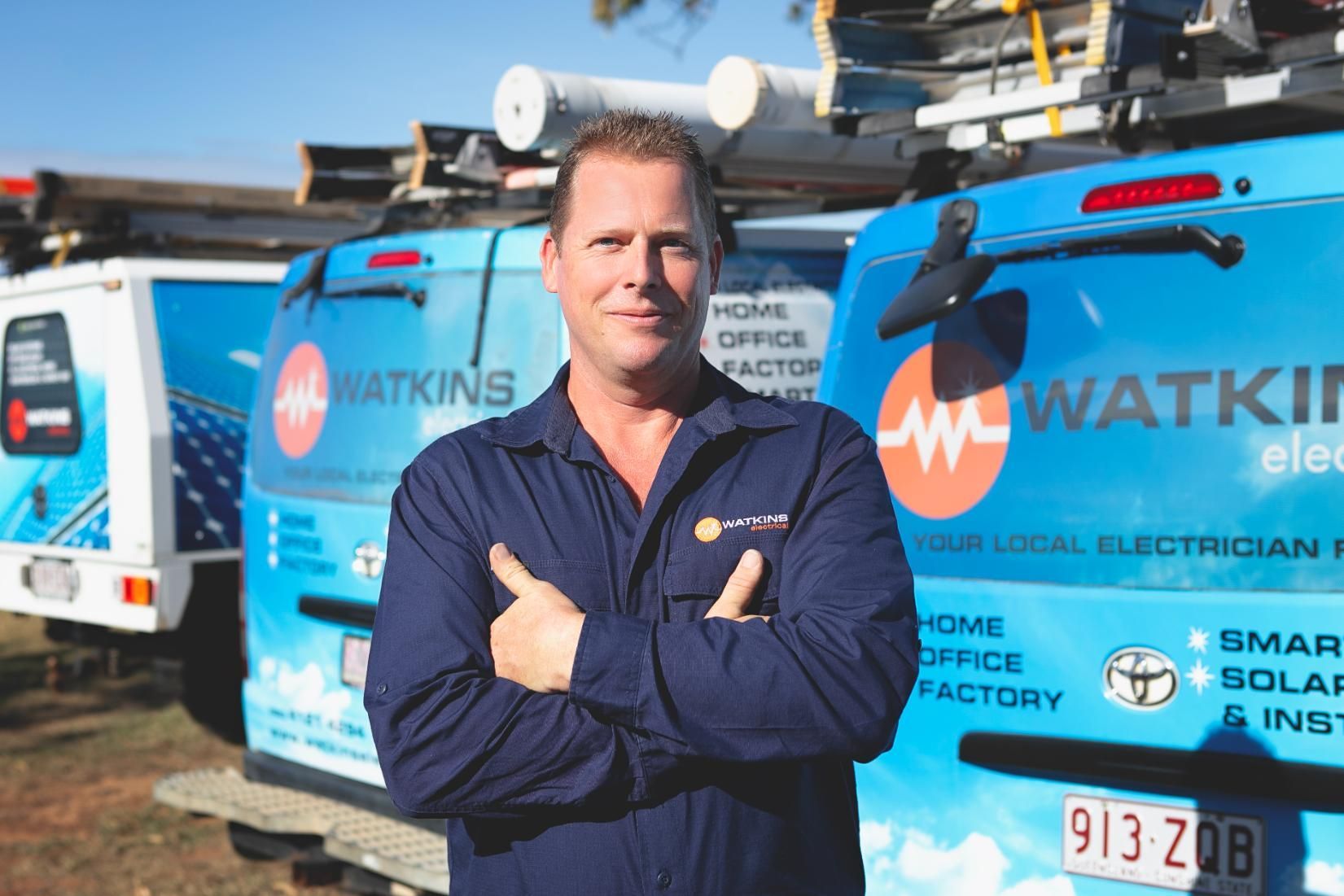 Man in blue uniform with arms crossed, standing in front of Watkins Electrical vans — Watkins Electrical In Pialba, QLD