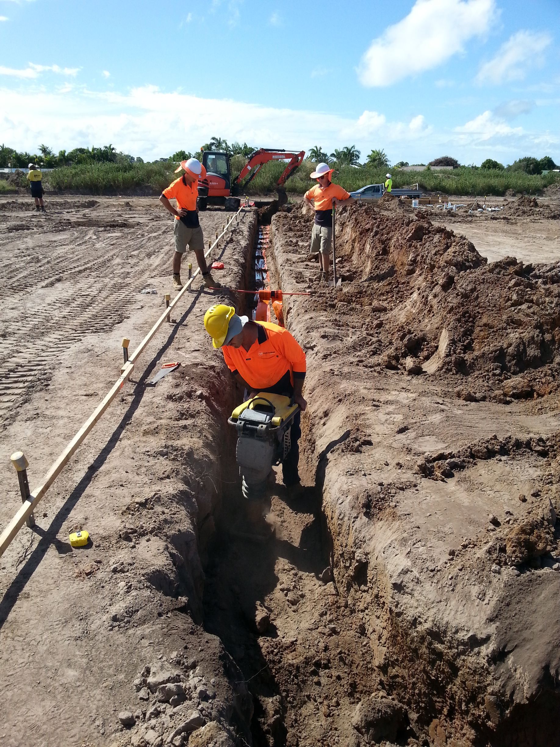 Construction workers in orange vests and hard hats operating machinery in a trench. Sunny, outdoor setting — Watkins Electrical In Pialba, QLD