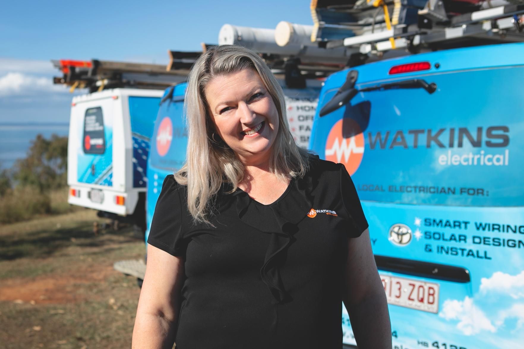 Woman in black shirt smiling in front of Watkins Electrical vans — Watkins Electrical In Pialba, QLD
