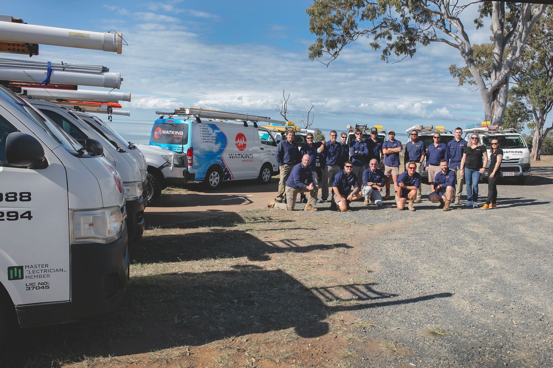 Group of workers in blue shirts pose with vans in a gravel lot — Watkins Electrical In Pialba, QLD