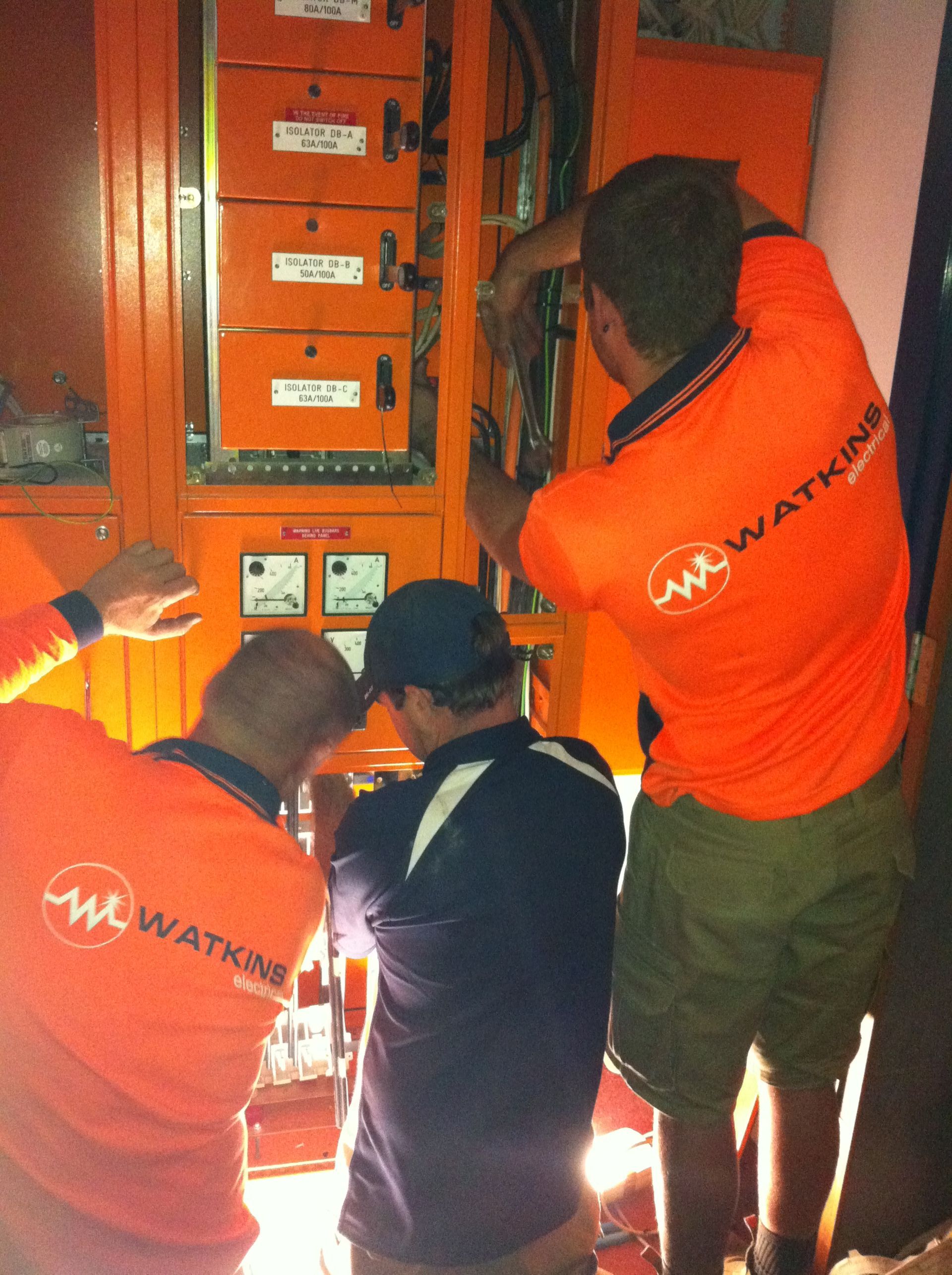 Three electricians in orange shirts work on an orange electrical panel — Watkins Electrical In Pialba, QLD