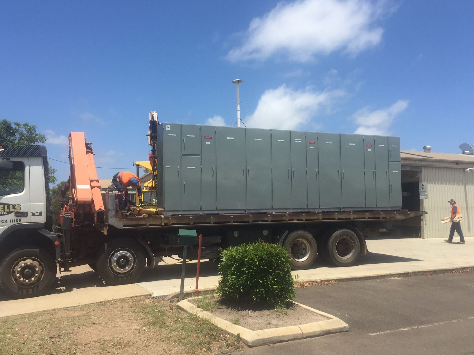 A large grey electrical unit on a flatbed truck, being unloaded by workers. Sunny day — Watkins Electrical In Pialba, QLD