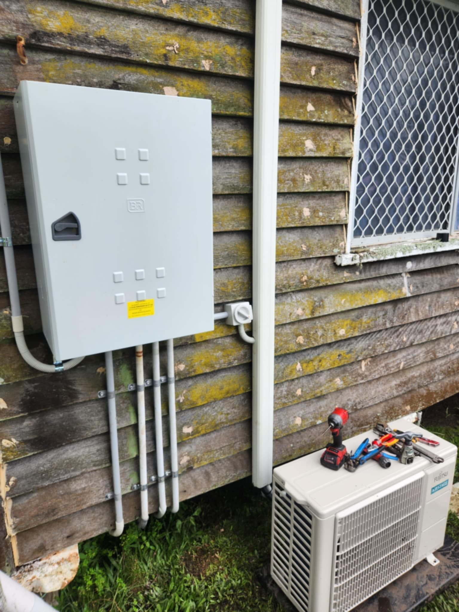 Air conditioner unit and electrical box on a weathered wooden building, with tools resting on the unit — Watkins Electrical In Pialba, QLD