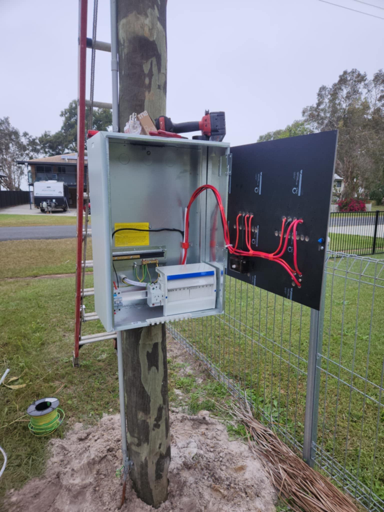 Electrical box on a utility pole with open door, tools, and wiring, in a yard setting — Watkins Electrical In Pialba, QLD