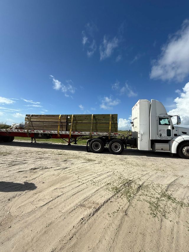 White flatbed truck hauling lumber on a sandy lot under a bright blue sky