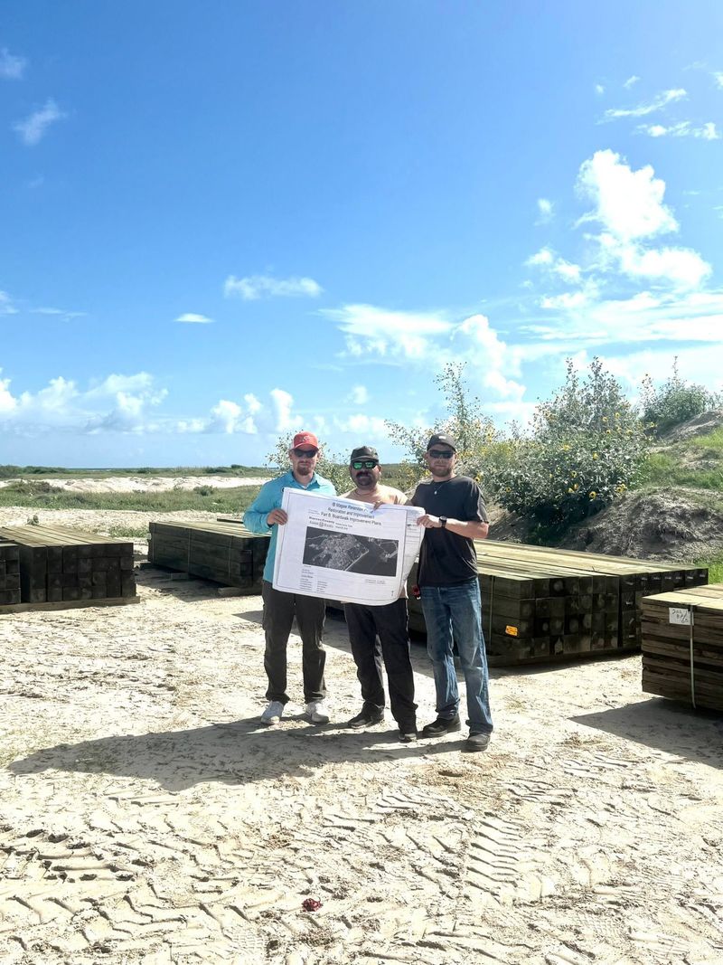 Three people holding a sign on a stone patio with the ocean and blue sky in the background.