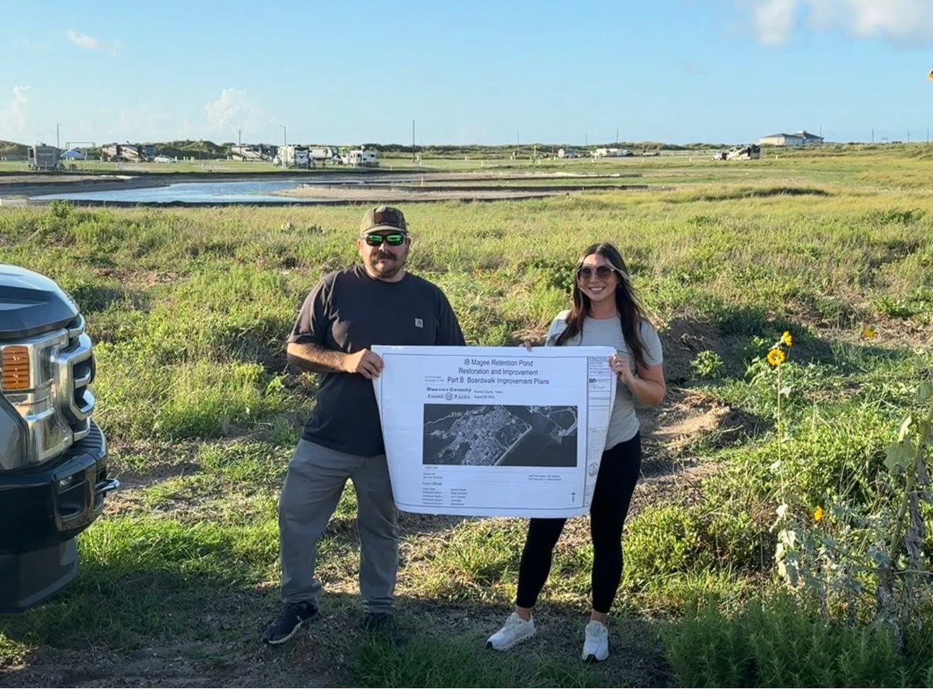 Two people holding a large poster beside a road in a grassy field on a sunny day