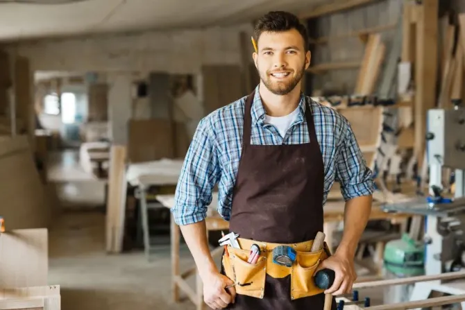 A smiling woodworker in a blue plaid shirt and brown apron with a tool belt, standing in a busy carpentry workshop.