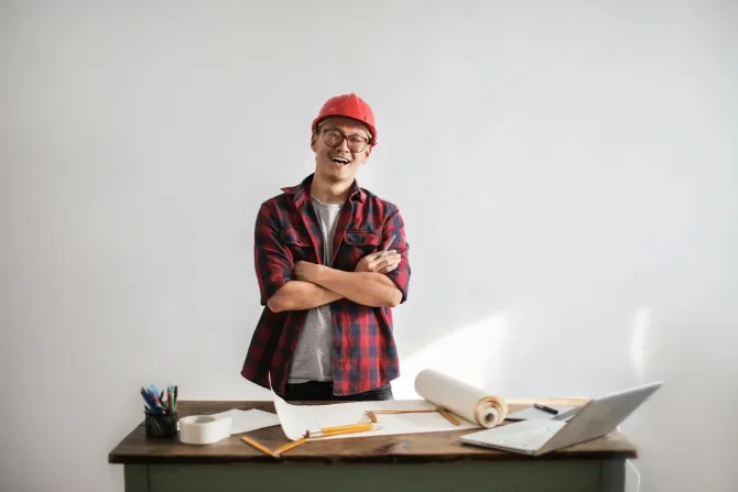 A person in a red hard hat and plaid shirt smiling with arms crossed, standing behind a desk with blueprints and a laptop.