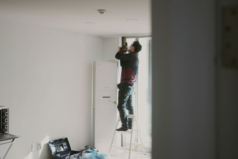 A person stands on a step ladder to inspect a ceiling fixture inside a white, sparsely furnished room.