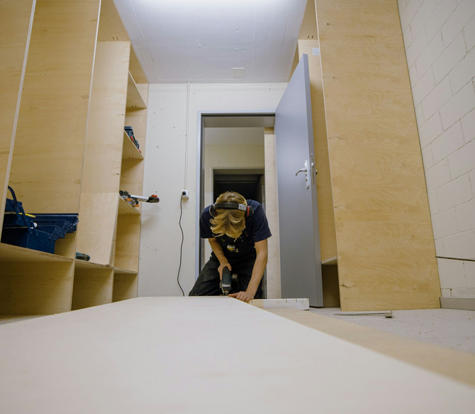 A person in a workshop uses a drill on a wooden panel, positioned between two floor-to-ceiling plywood storage units.