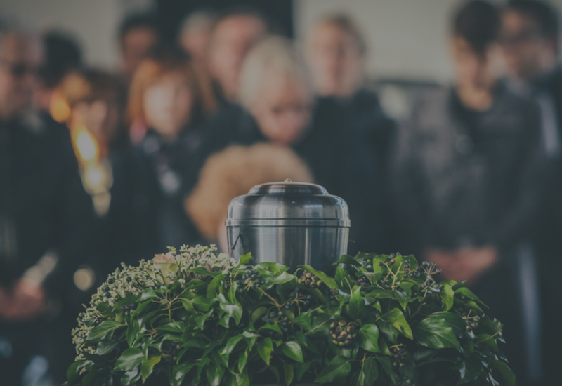 Black urn on a leafy memorial display with blurred mourners in the background