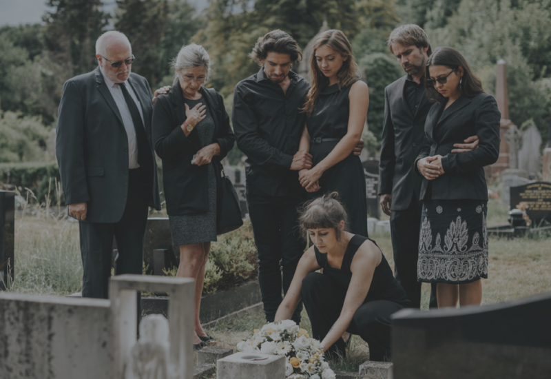 Mourners stand around a gravesite as a woman places flowers on a grave marker.