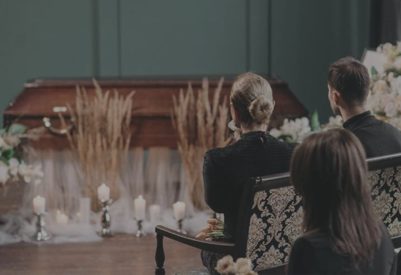 Audience seated in a dim hall, facing a coffin surrounded by candles and flowers