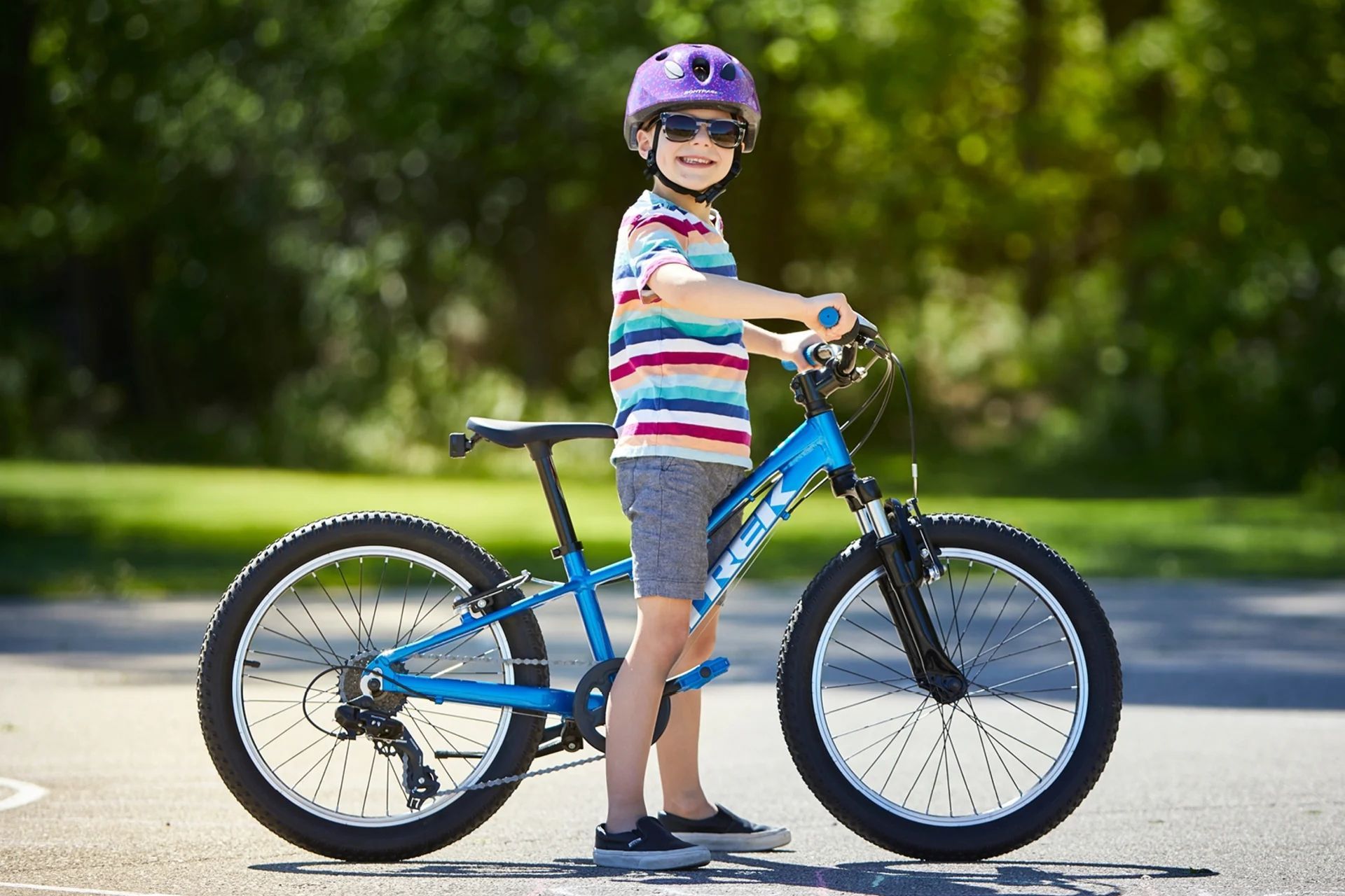 Een jonge jongen met een helm en een zonnebril staat naast een blauwe fiets.
