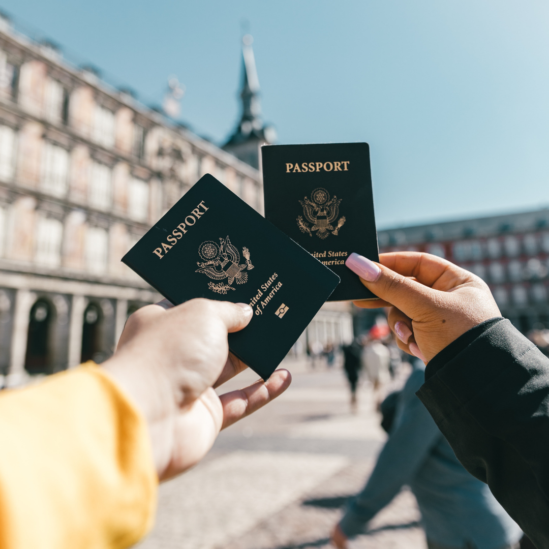 Two travelers holding U.S. passports in a European plaza on a sunny day.

