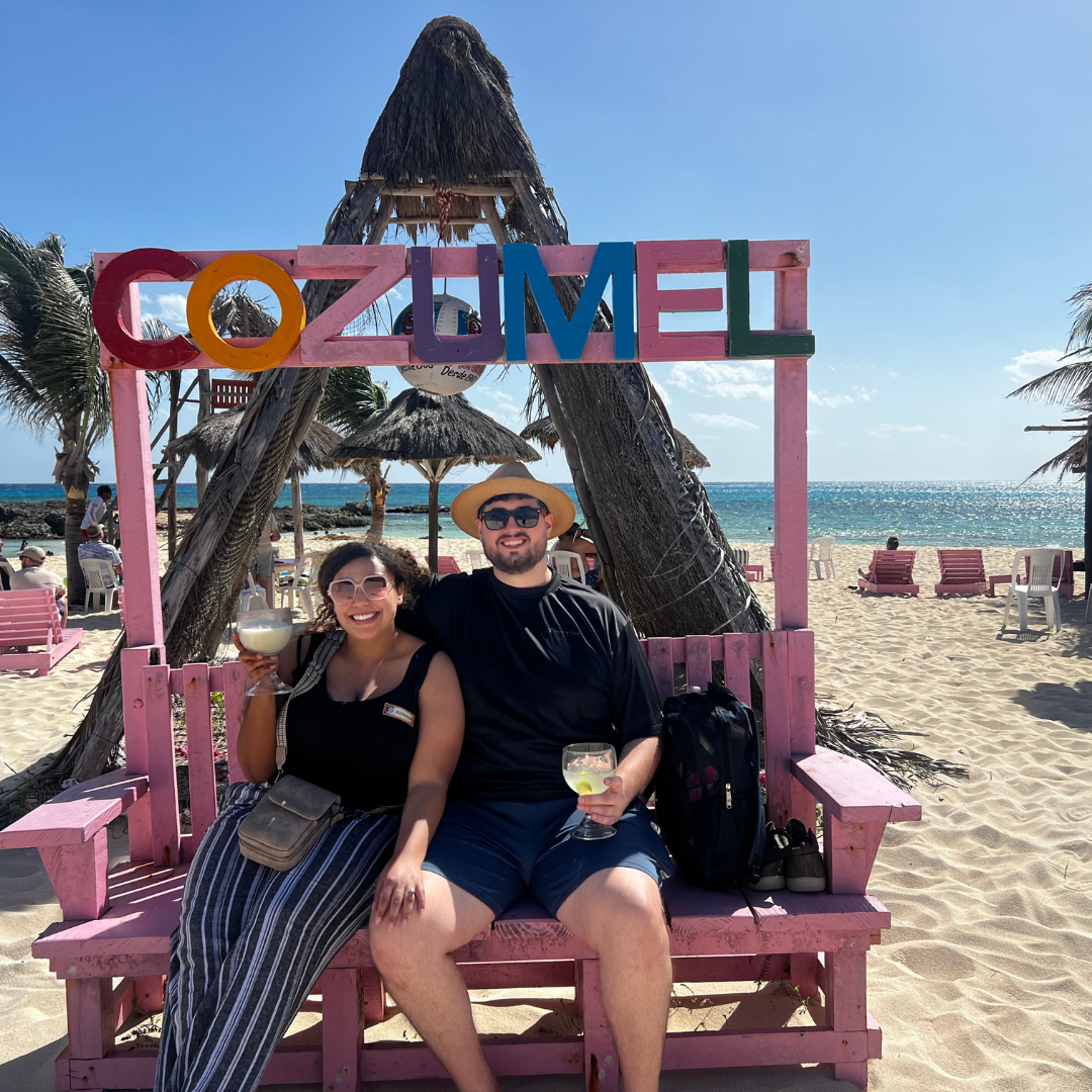 Couple with tropical drinks under a colorful “Cozumel” sign on a beach in Mexico.
