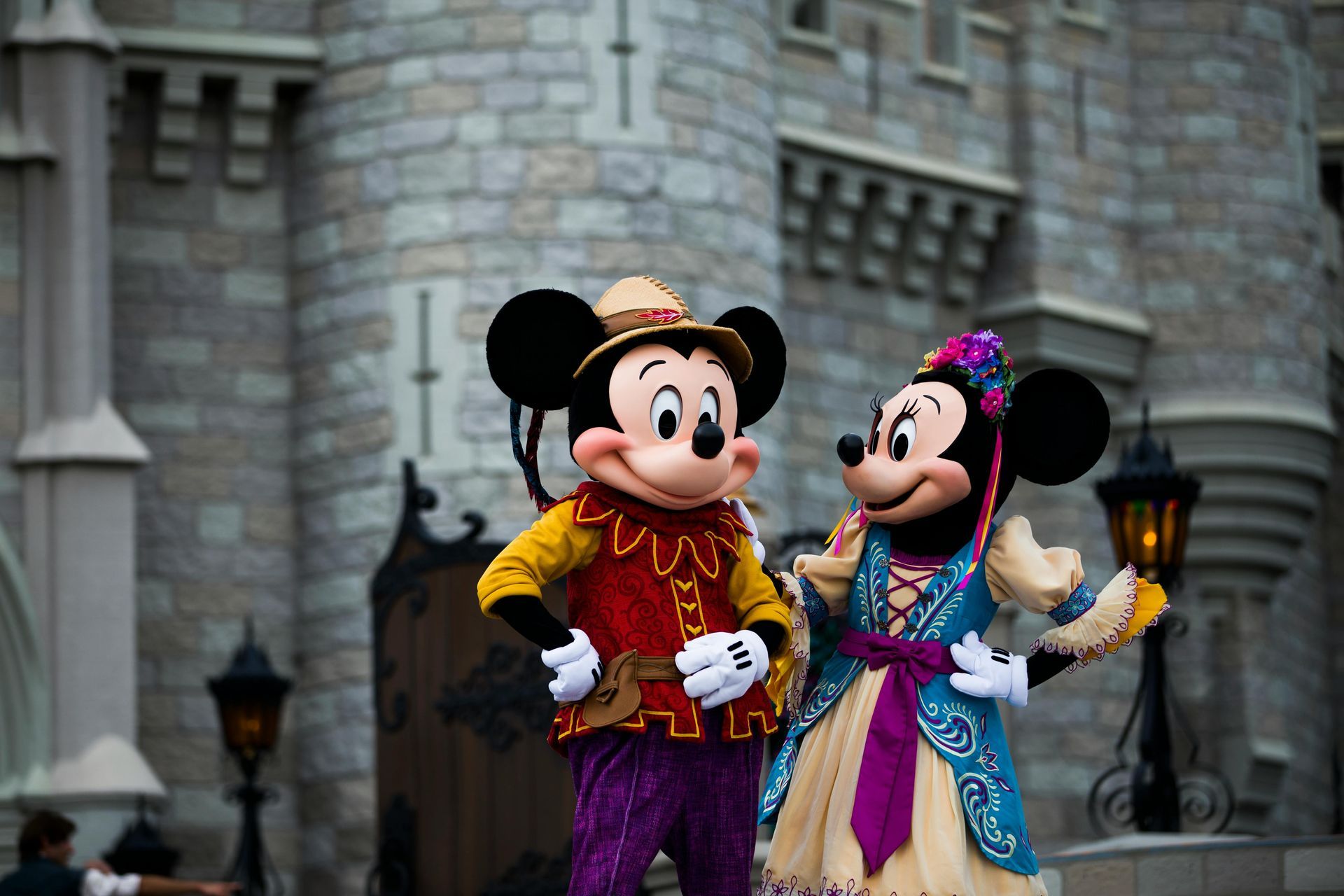 Mickey and Minnie Mouse dressed in festive costumes greeting guests at a Disney theme park in front of a castle backdrop.