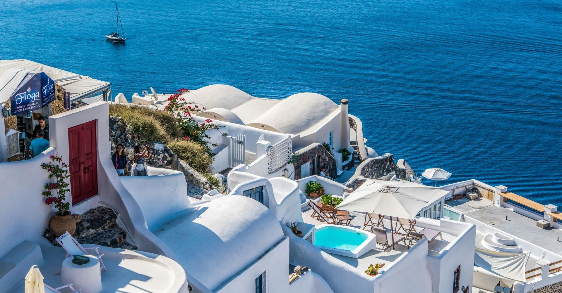 Cliffside villas in Oia, Santorini overlooking the Aegean Sea with a sailboat and plunge pools.
