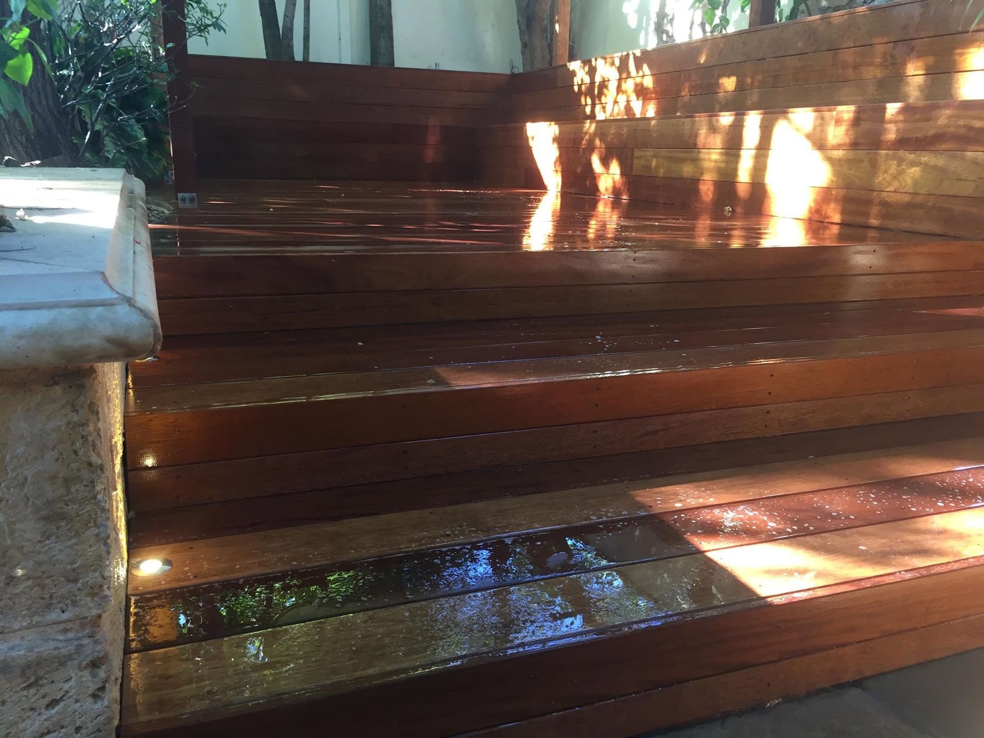 Wooden outdoor benches in dappled sunlight, with wet steps and a reflective puddle nearby.