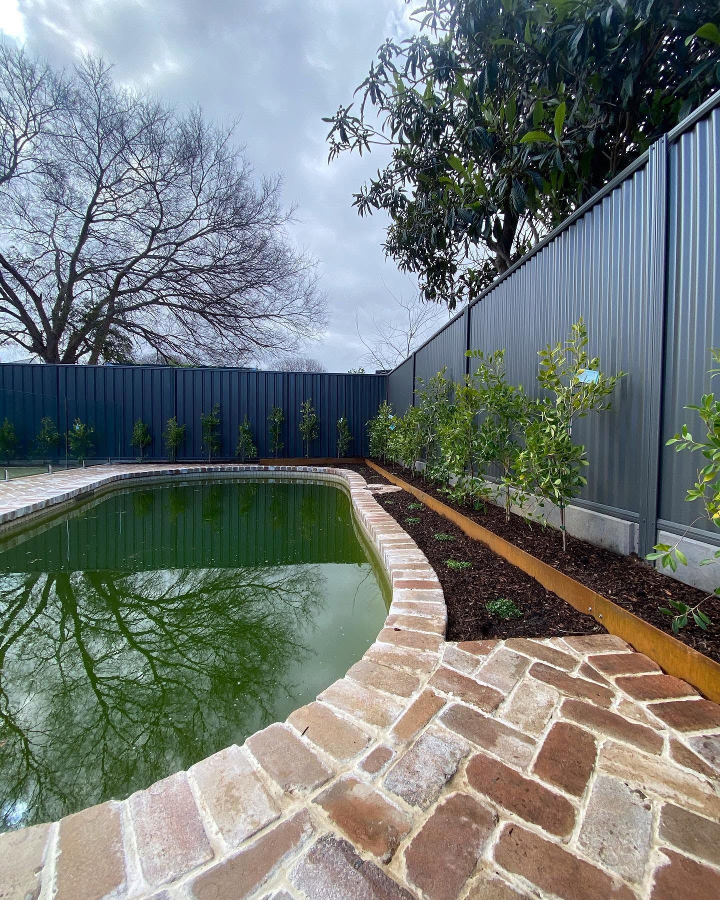 Backyard pool with stone patio, metal fence, and greenery along the edge