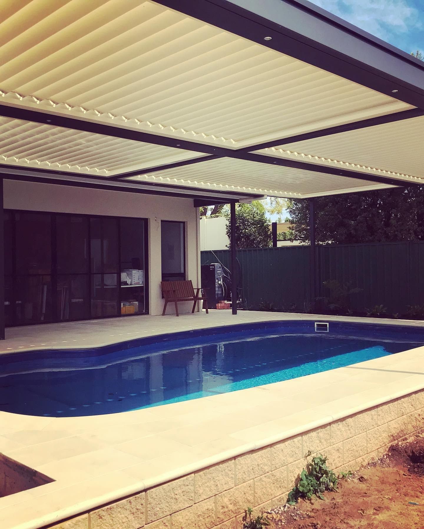 Covered backyard swimming pool beside a patio with chairs and a dark fence