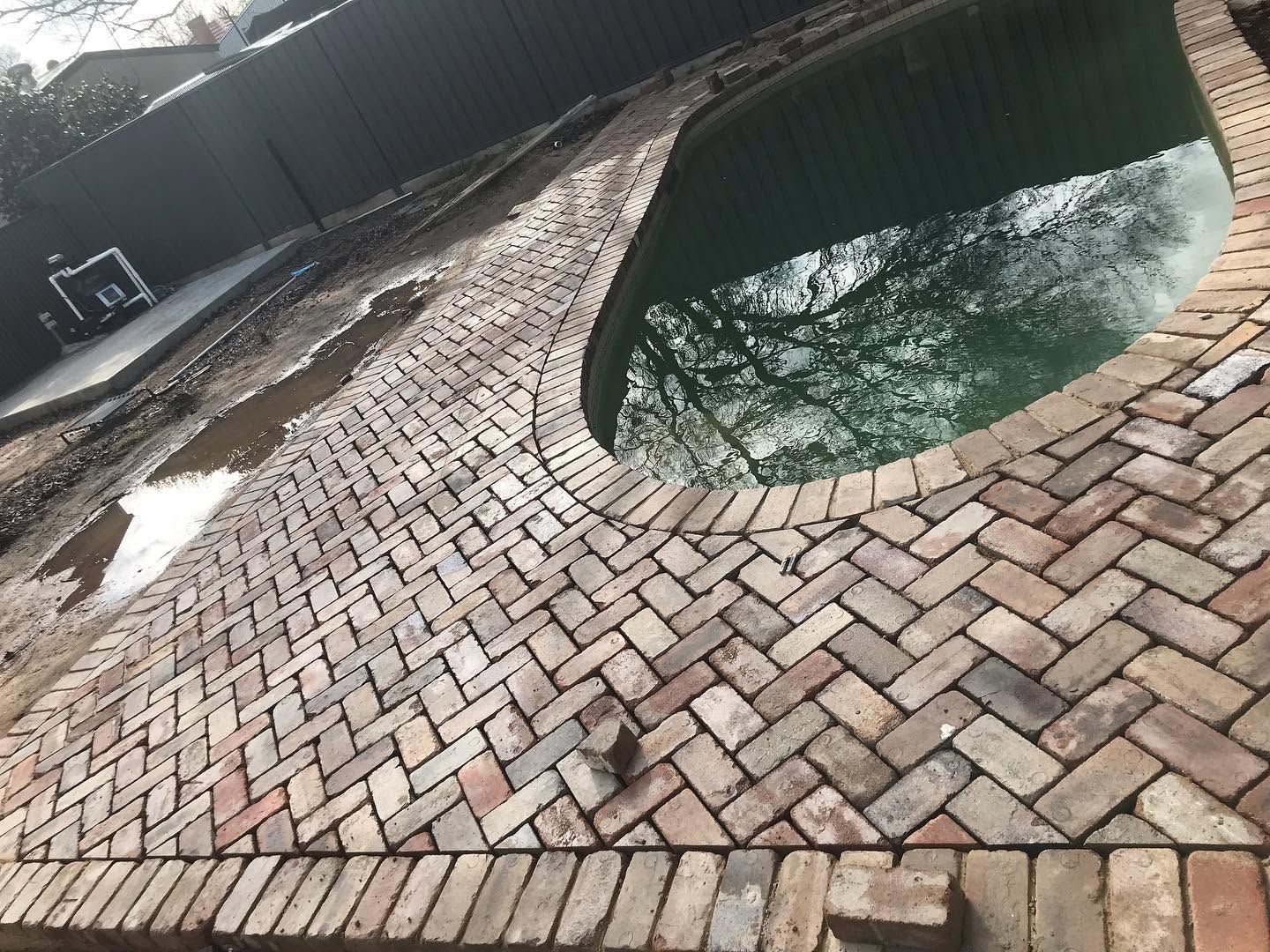 Brick pool deck beside a dark green swimming pool, viewed at a slight angle.