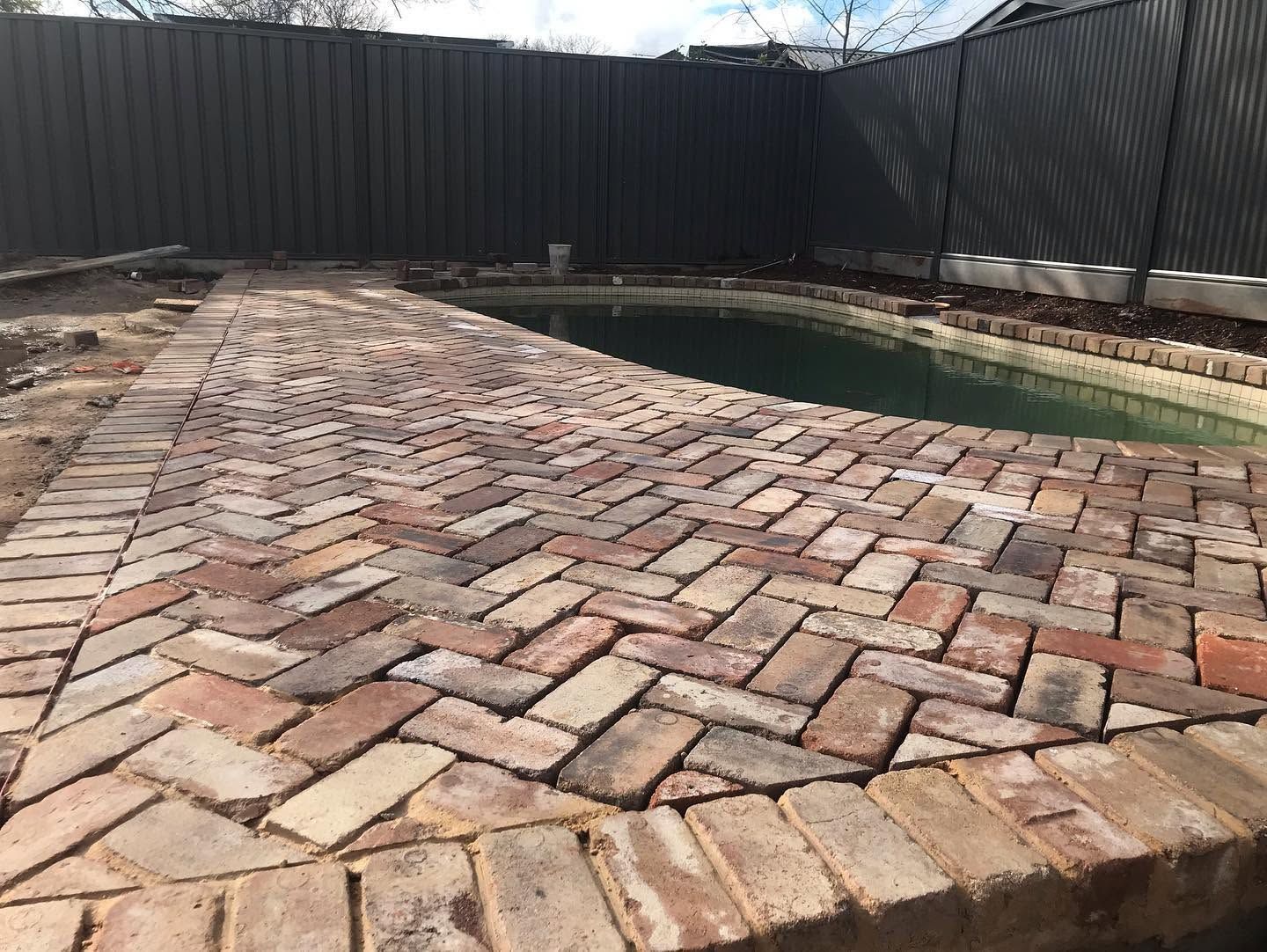 Brick patio beside a narrow swimming pool, bordered by a black fence.