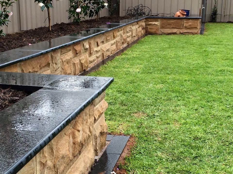 Curved stone retaining wall with dark coping borders a green lawn in a landscaped backyard.