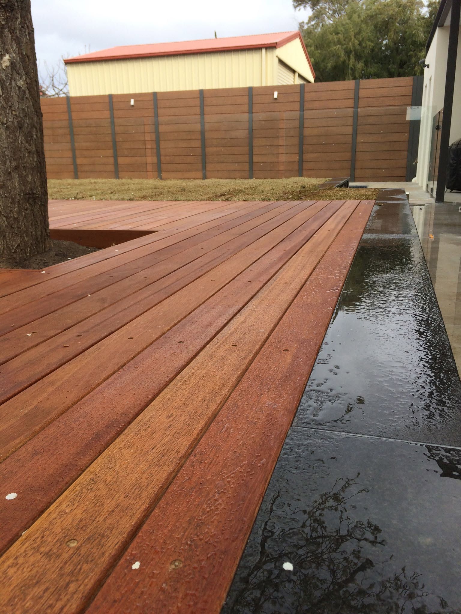 Red wooden planks laid on wet pavement beside a backyard fence and house.