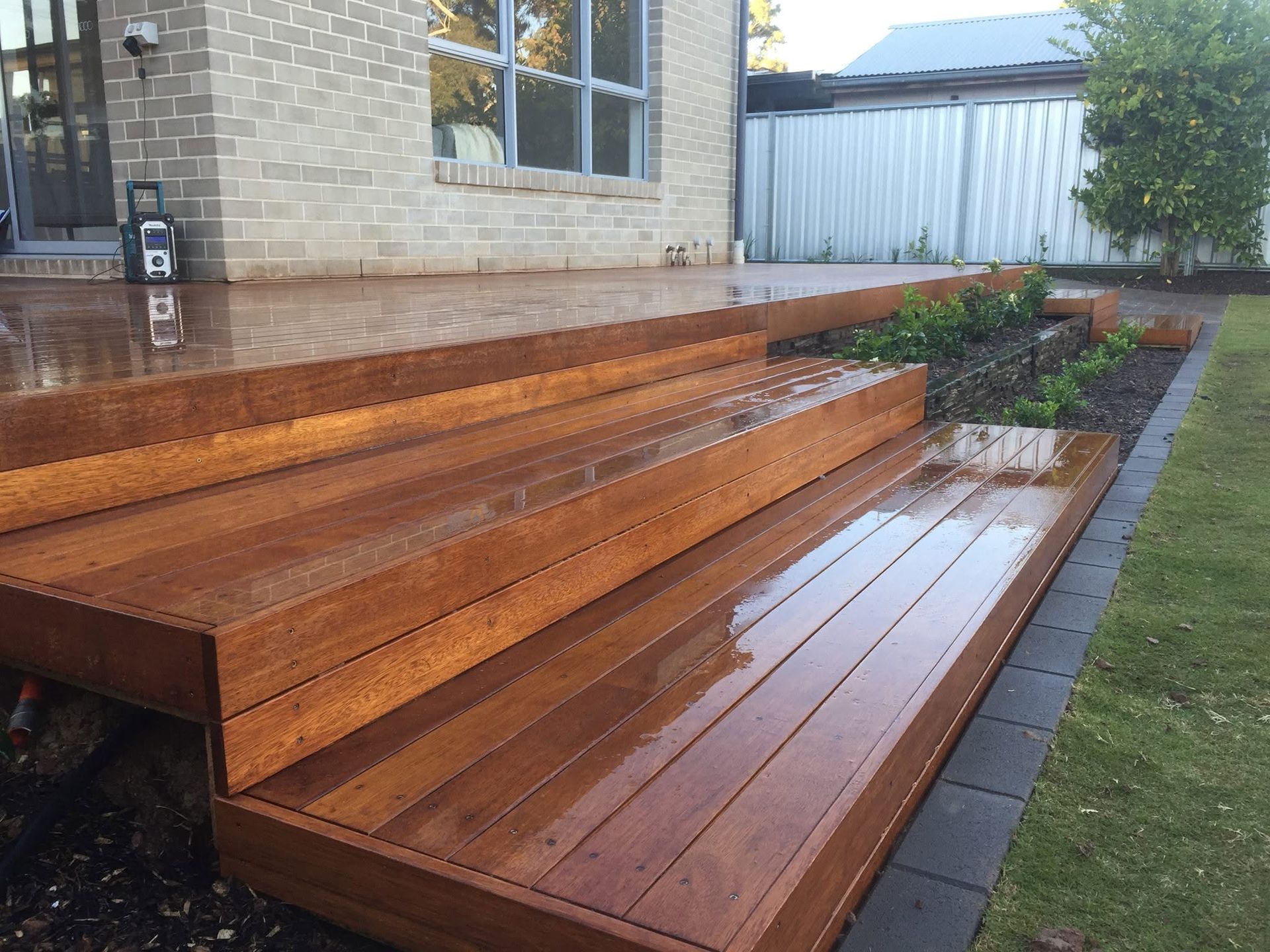 Wet wooden deck steps beside a house, with rainwater reflecting on the boards and a garden bed nearby