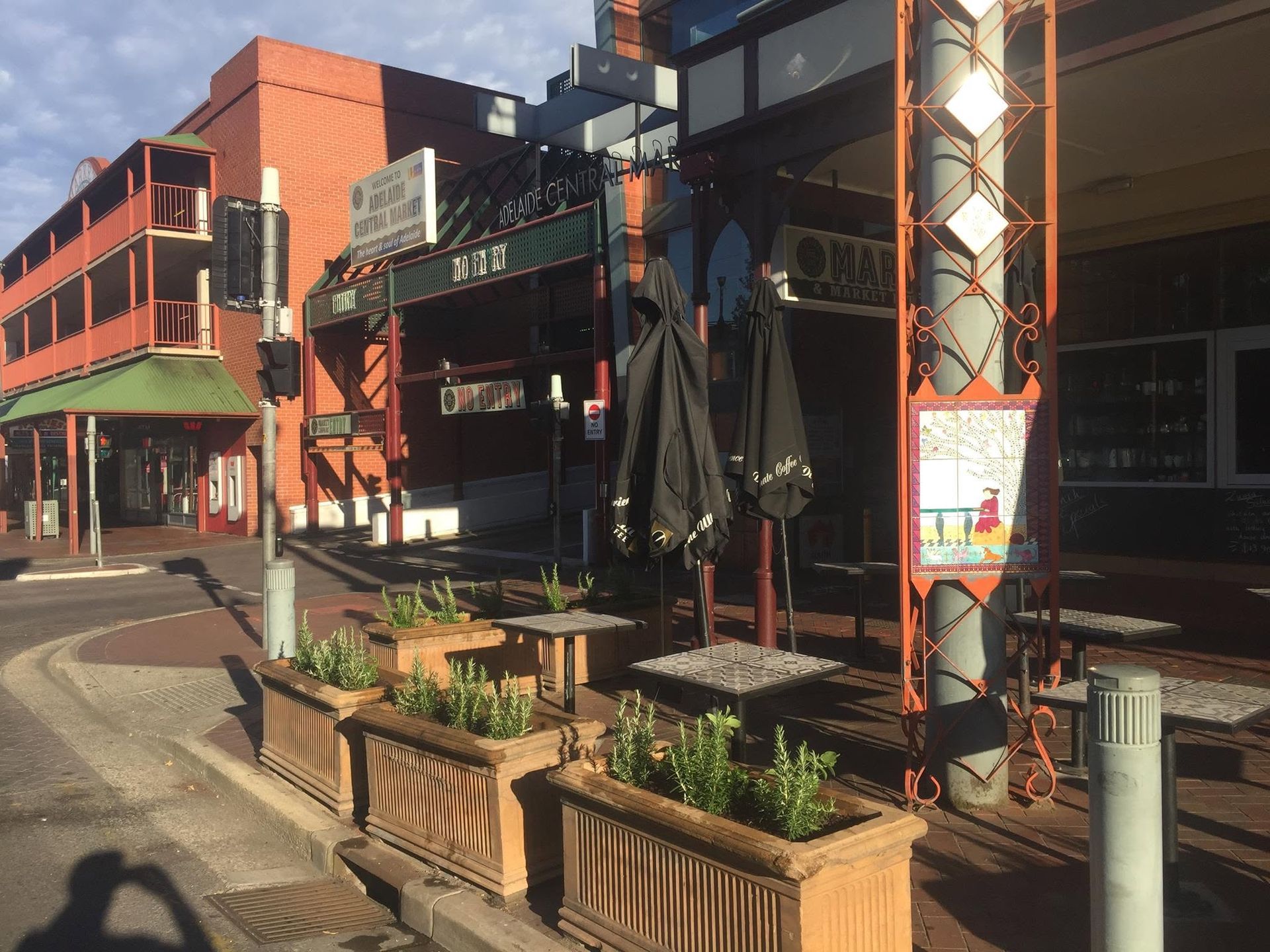 Sunny street with brick buildings, storefronts, and outdoor planters along a curved sidewalk