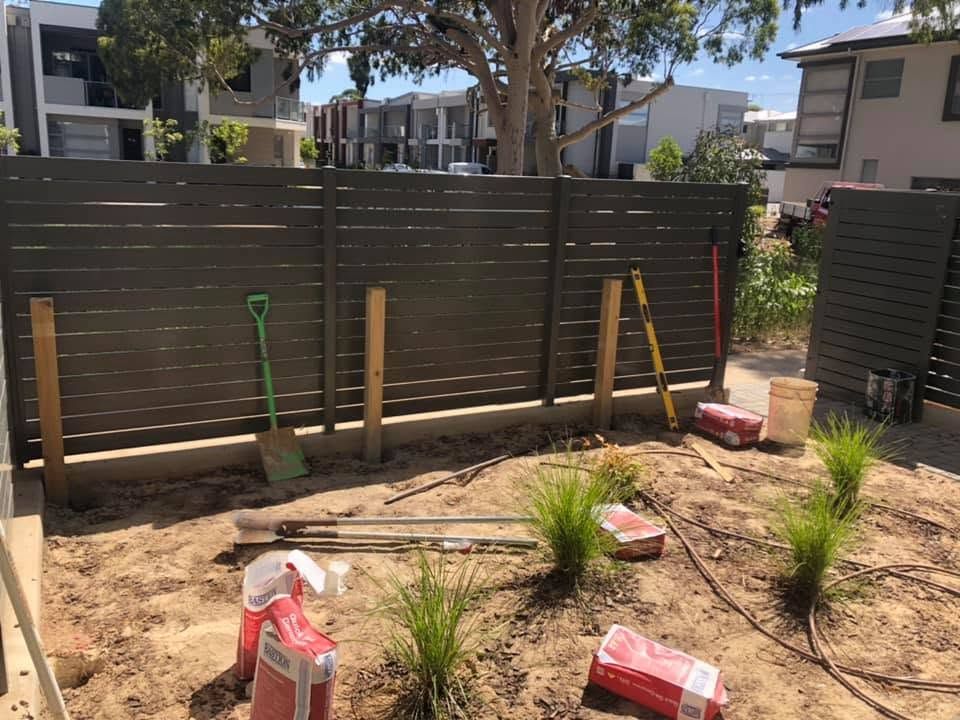 Backyard garden bed with new plants, shovels, and a dark wooden fence under construction