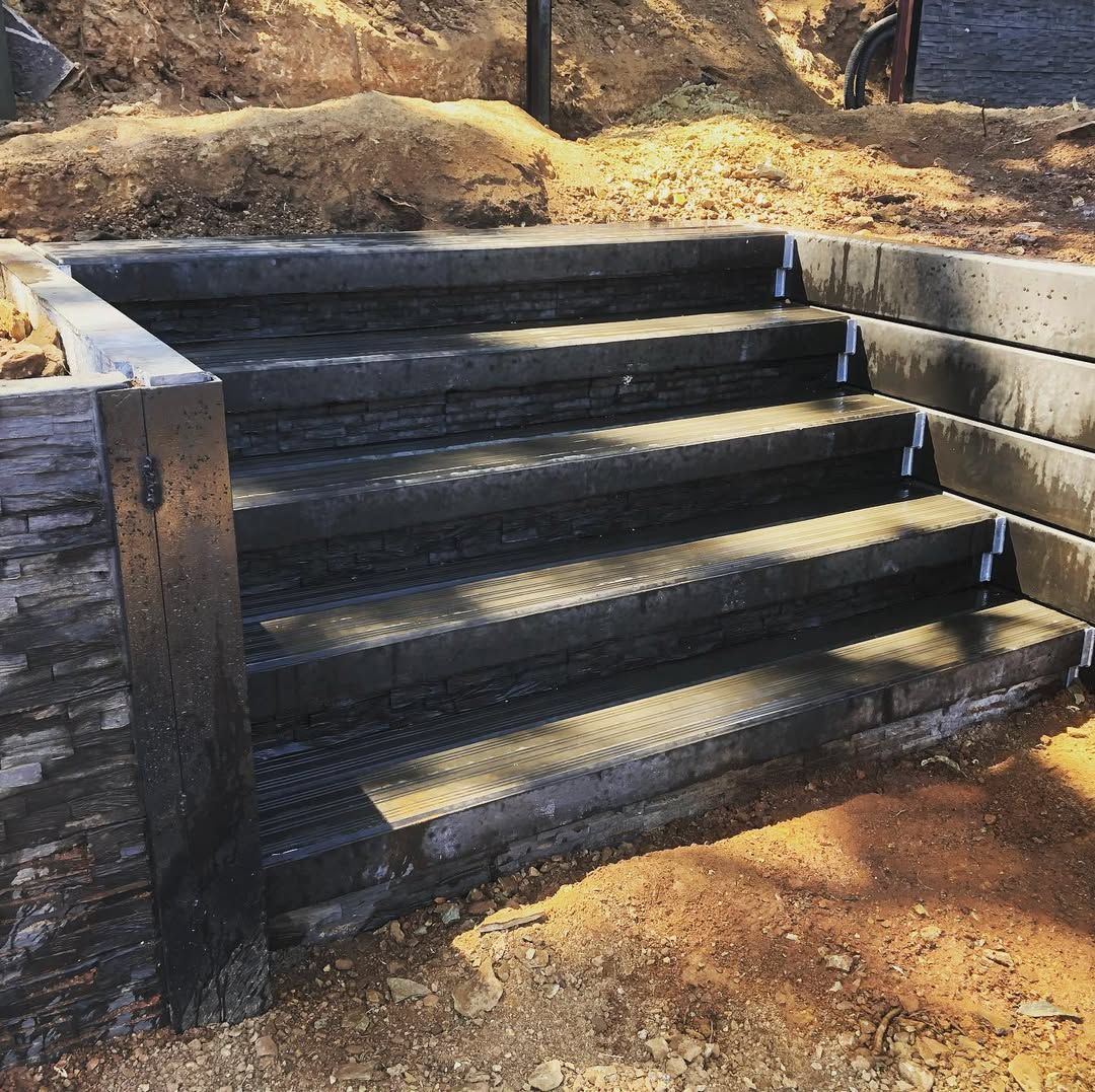 Dark stone steps with black railings leading up a dirt embankment in a wooded area.