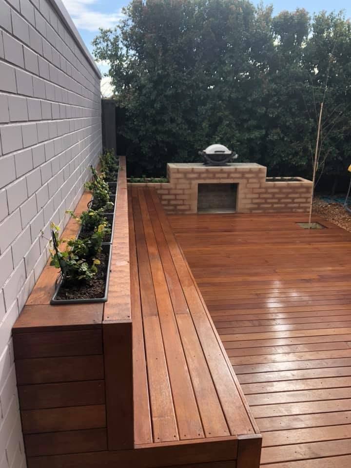 Wooden deck with built-in planter along a wall, leading to a brick outdoor grill area surrounded by trees.