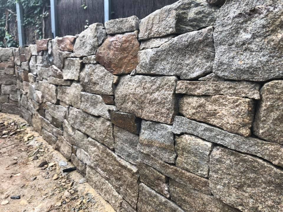 Stone retaining wall along a dirt path beside a fence, made of stacked rough-cut blocks.