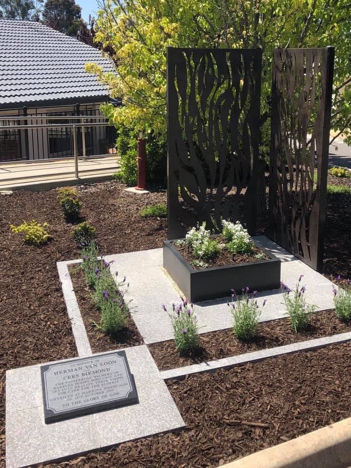 Outdoor memorial garden with a black metal sculpture, plaques, and planted beds beside a building. Outdoor memorial garden with a black metal sculpture, plaques, and planted beds beside a building.